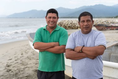 İki gülümseyen balıkçı kumlu bir sahilde arka planda küçük tekneleri ve dağları olan