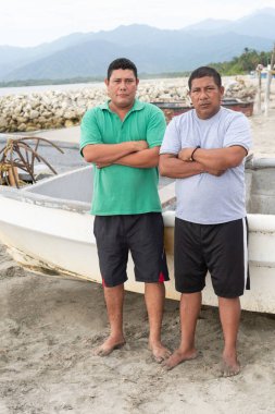 Çıplak ayakla duran balıkçılar kumlu bir sahilde kollarını kavuşturmuş bir teknenin yanında.