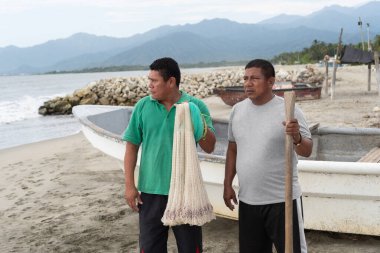 İki Latin Amerikalı balıkçı, bir balık ağı ve tekneyle sahilde dikiliyorlar.