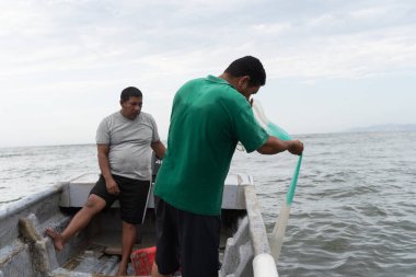 Küçük bir teknedeki balıkçılar bulutlu bir gökyüzünün altında denizden balık ağı çekiyorlar.