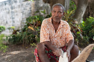 Yaşlı bir adam bir aletle ahşap oyuyor, tropik bir ortamda sanatına odaklanıyor.