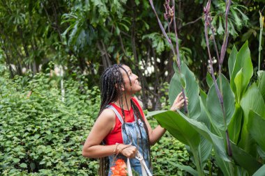 Bir kadın bir bitkiye dokunuyor, yeşil bahçe ortamında merak ve neşe yaşıyor.