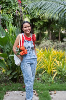 Parlak bir şekilde gülümseyen, çantasındaki turuncu gülleri taşıyan, doğadaki yavaş verimliliği kucaklayan bir kadın.