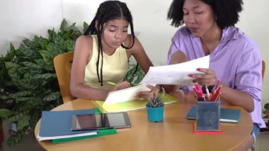 Afrikalı Amerikalı anne genç kızına ev ödevlerinde yardım ediyor, eğitici materyalleri anlatıyor.