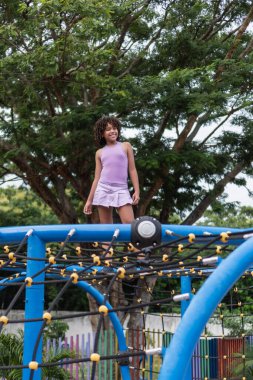 Rastalı genç bir kız oyun parkında dikiliyor ve gülümsüyor.