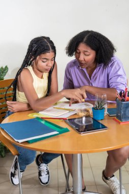 Yetişkin bir kadın, genç bir kıza ahşap masada ders veriyor.