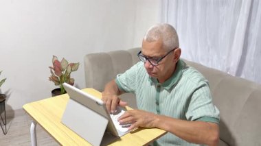 Odaklanmış olgun bir adam koltukta oturuyor ve oturma odasındaki modern tablet üzerinde çalışıyor.