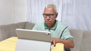 Gözlüklü yaşlı bir adam oturma odasından çalışırken tablet ekranına konsantre oluyor.