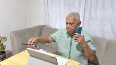 Gözlüklü yaşlı adam bir tablet üzerinde çalışırken akıllı telefonuna cevap veriyor.