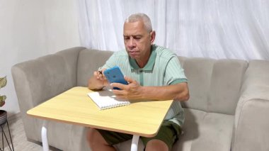Kanepede oturan yaşlı bir adam, cep telefonu kullanıyor ve oturma odasında notlar yazıyor.