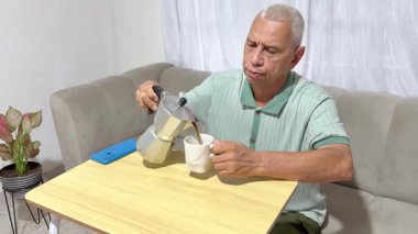 Olgun bir adam kanepede oturup günlük rutininin tadını çıkarıyor, sıcak kahveyi evdeki bir fincana dolduruyor.