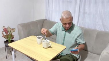 Olgun bir adam evdeki kanepede oturup telefonunu kontrol ederken kahvesini tatlandırıyor.