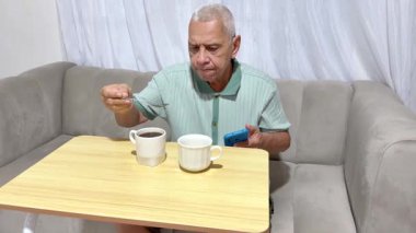 Kıdemli adam günlük rutininin tadını çıkarıyor, kahve içiyor ve kanepede akıllı telefonuna bakıyor.