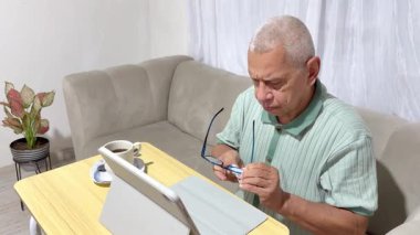 Kahvesini içmeden ve tablet üzerinde çalışmadan önce gözlüğünü temizleyen gri saçlı son sınıf öğrencisi.