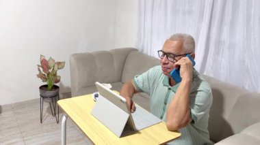 Gözlüklü yaşlı adam telefonda konuşuyor ve kanepede kahve içerken tablet kullanıyor.