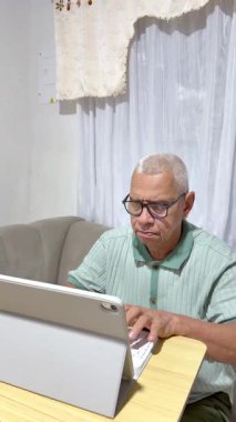 Gözlüklü yaşlı adam tablet klavyeye yazarken konsantre oluyor, evden çalışıyor.