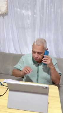 Olgun bir adam ciddi bir telefon görüşmesi yapıyor ve evdeki kanepede otururken tablet üzerinde çalışıyor.