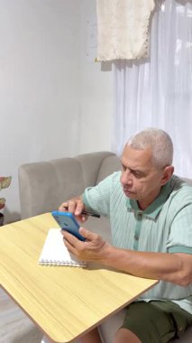 Yaşlı adam akıllı telefonu özenle kullanıyor ve bütçe yönetimi için not tutuyor.