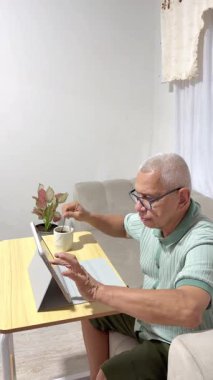 Yaşlı bir adam oturma odasında bir fincan sıcak kahve içerken tablet bilgisayarda uzaktan çalışıyor.