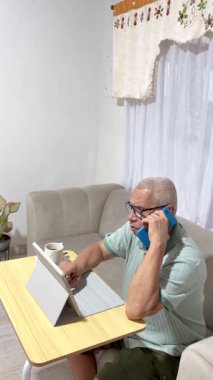 Olgun bir adam bir tablet üzerinde çalışıyor ve oturma odasında telefonla konuşuyor, uzaktan kumandayı kucaklıyor.