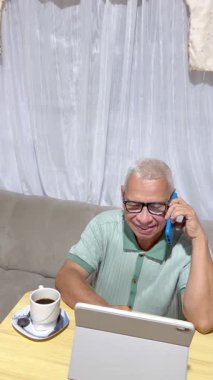 Olgun adam birden fazla iş yapıyor, telefonda konuşuyor ve evde tablet üzerinde çalışırken kahve içiyor.