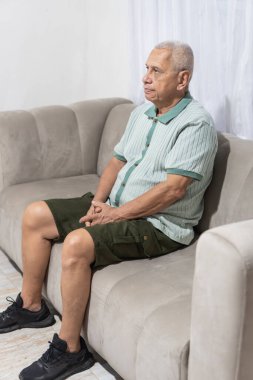 Evdeki kanepede tek başına oturan kıdemli adam, düşünceli görünüyor.