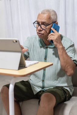 Kıdemli adam birden fazla iş yapıyor, evde tablet kullanırken telefonla konuşuyor.