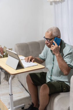 Evden uzakta çalışan kıdemli bir adam tablet yazarken telefonla konuşuyor.