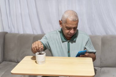 Kanepede oturan kıdemli bir adam akıllı telefon kullanıyor ve portatif bir masada kahve karıştırıyor.