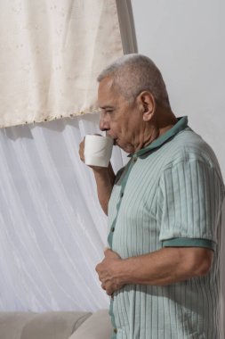 Yaşlı adam bardaktan su içiyor, sabah ayini sırasında pencereyi içeriden gözetliyor.