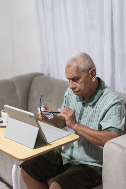 Üst düzey bir adam gözlüğünü temizlerken, evde dijital tablet kullanmaya hazırlanıyor.