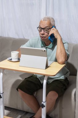 Kanepede oturan kıdemli bir adam, tabletle birden fazla iş yapıyor ve akıllı telefonuyla konuşuyor.