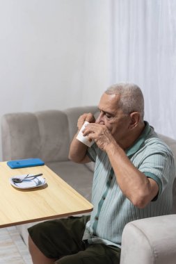 Yaşlı adam kanepede oturuyor, evde dinlenirken bir bardaktan içiyor.
