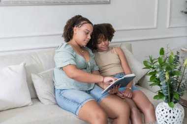 İki kız kardeş kanepede oturuyor, birlikte kitap okuyor, öğrenme anını paylaşıyorlar.