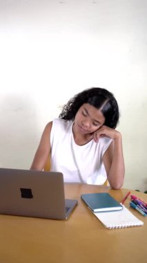 Masasında dizüstü bilgisayar ve kitapla çalışırken esneyen ve uykusuzlukla savaşan Asyalı genç bir kadın.