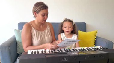 Genç anne ve küçük kızı nota çalışıyor ve piyano öğrenirken birlikte şarkı söylüyorlar.
