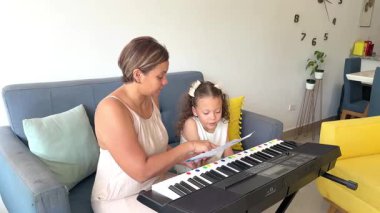 Genç bir kız annesiyle özel müzik dersi alıyor ve oturma odasında elektrikli piyano çalıyor.