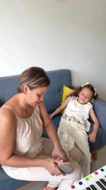 Şefkatli anne, gülümseyen kızının ayakkabılarını giymesine yardım ederken evdeki mavi koltukta oturuyor.