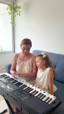 Bayan öğretmen küçük bir kız öğrenciye oturma odasında elektronik piyano çalmayı öğretiyor.
