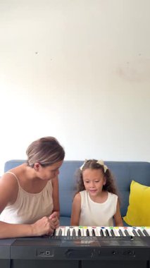 Genç bir kız annesiyle birlikte oturma odasında piyano dersi alıyor, müzik becerileri geliştiriyor.