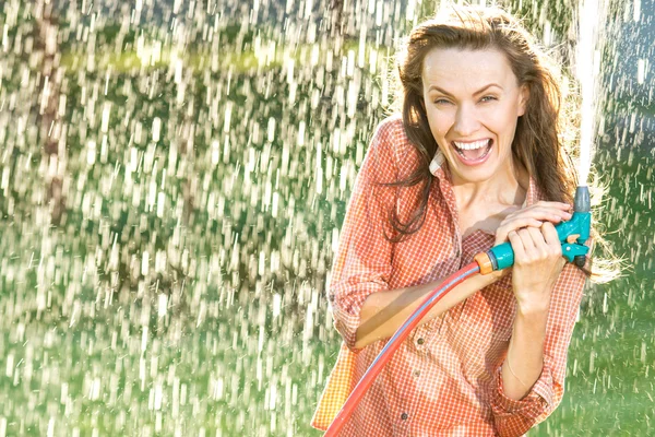 Hermosa mujer joven que se divierte en el jardín de verano con manguera de jardín salpicadura lluvia de verano —  Fotos de Stock