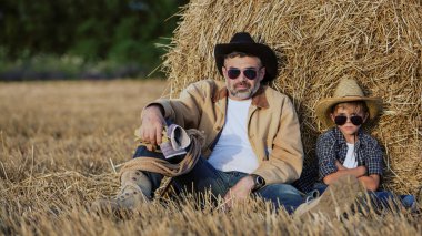 Baba ve oğlu tarlada dinlenirken şapka, gömlek ve kot pantolon giyerler. Evlat, baba kavramı gibi.