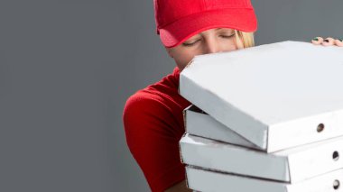 Kırmızı şapkalı, boş tişörtlü bir kadın servis elemanı yemek siparişlerini gri arka plan stüdyosunda izole edilmiş pizza kutularına veriyor.