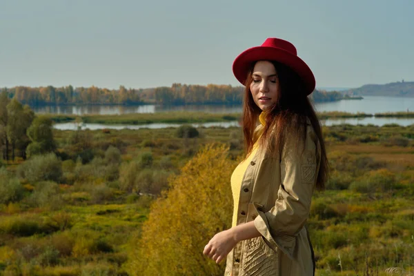 Дівчина в жовтому светрі плащ і червоний капелюх стоїть на тлі річки в осінньому пейзажі один — стокове фото