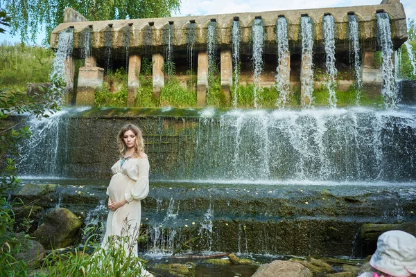 Молода красива вагітна дівчина в білій сукні і корона стоїть на тлі водоспаду — стокове фото