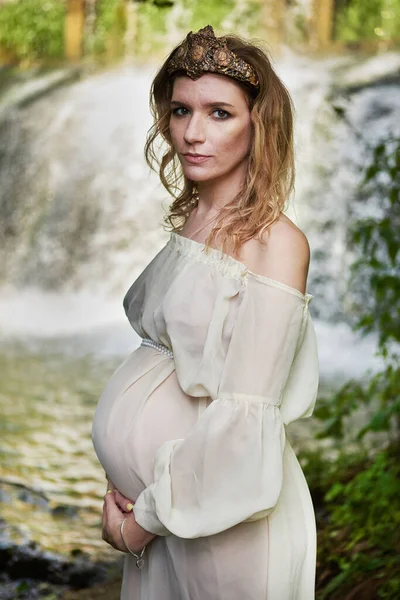 Молодая красивая беременная девушка в белом платье и корона стоит на фоне водопада — стоковое фото