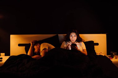 Geceleri yatakta akıllı telefonları olan genç bir çift.