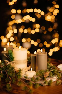 Bokeh ışıklarıyla samimi mum düzenlemesi. Karmaşık sütun mumlarından, çam dallarından ve kırsal ahşap masadaki yeşil aksanlardan gelen yumuşak parıltı, ılık festival atmosferi.,