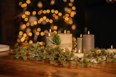 Kır masasında sıcak mum aranjmanı, gri ve krem mumlar, çam dalları ve koniler, altın renkli yumuşak alevler, rahat kızlık zarı atmosferi,