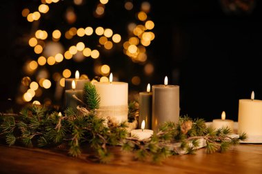 Ahşap masaya yayılmış şenlikli mum, çoklu alevler ve sonsuza kadar yeşil çelenk, sıcak altın bokeh ağacı, samimi ev sahipliği sahnesi, tatil yemeği hazırlığı.,